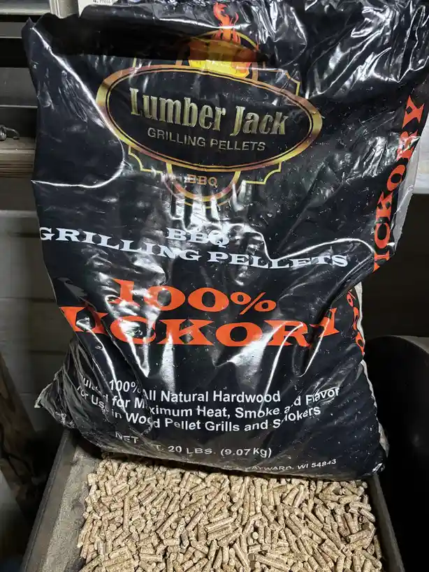 A large black bag of Lumber Jack brand BBQ grilling pellets, labeled '100% KICKOR', sits with its top open, spilling light…