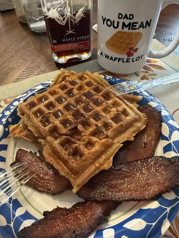 A close-up photo of a breakfast plate with two waffles topped with syrup and pecans, alongside several strips of crispy…