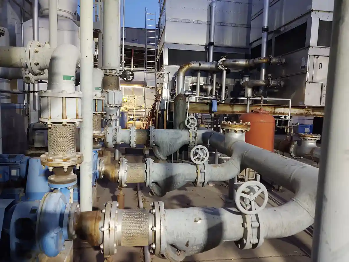 An outdoor industrial setting filled with large, rusted, interconnected pipes, valves, and machinery under bright sunlight…