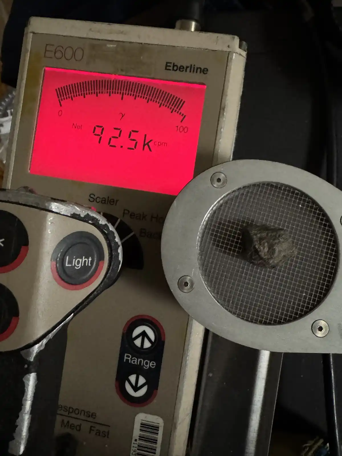 A close-up of an Eberline E600 radiation detector with a bright red display showing 'Net 92.5k cpm' while a small, rough…