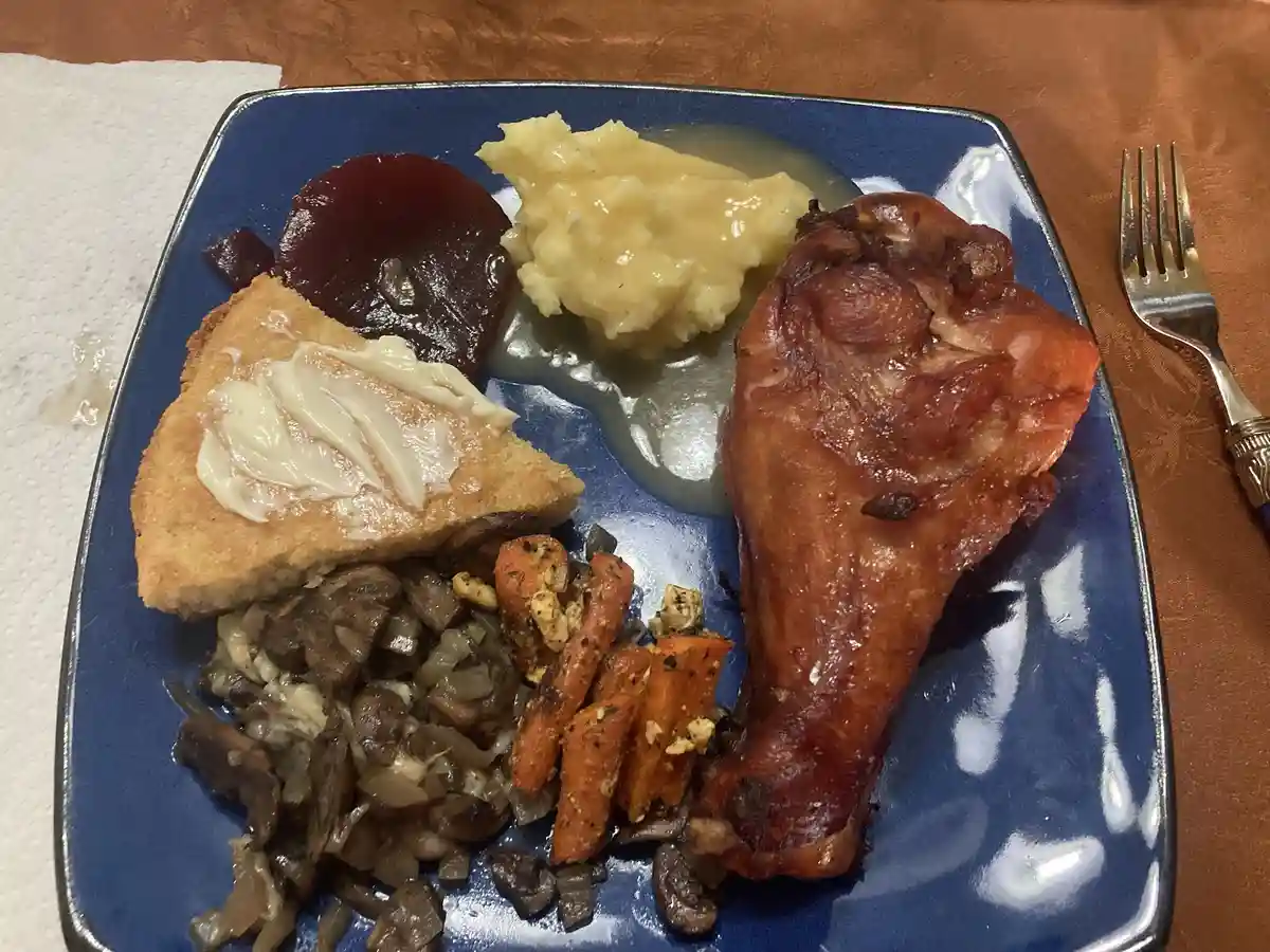 A blue plate holds a roasted chicken leg, mashed potatoes with gravy, cranberry sauce, buttered cornbread, sautéed mushrooms…