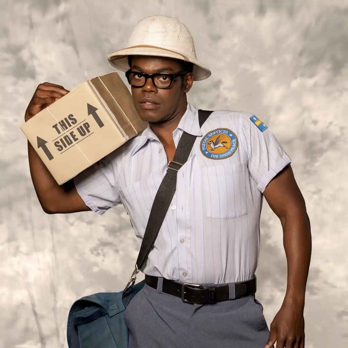 A man in a postal worker uniform with a pith helmet and glasses holds a cardboard box labeled 'THIS SIDE UP' while looking…