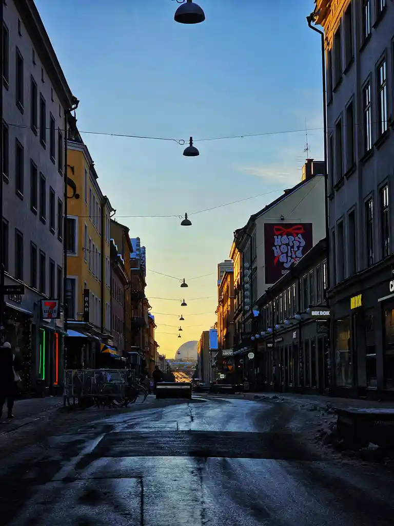 A narrow European street at dusk, lined with multi-story buildings, wet pavement reflecting the fading light, and hanging…