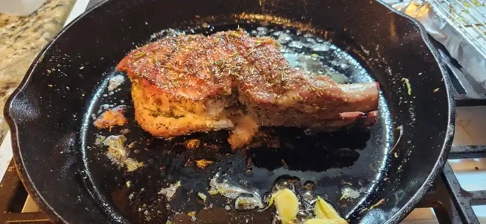 A close-up photo shows a large, seasoned steak sizzling in a black cast-iron skillet on a stovetop, with melted butter and…
