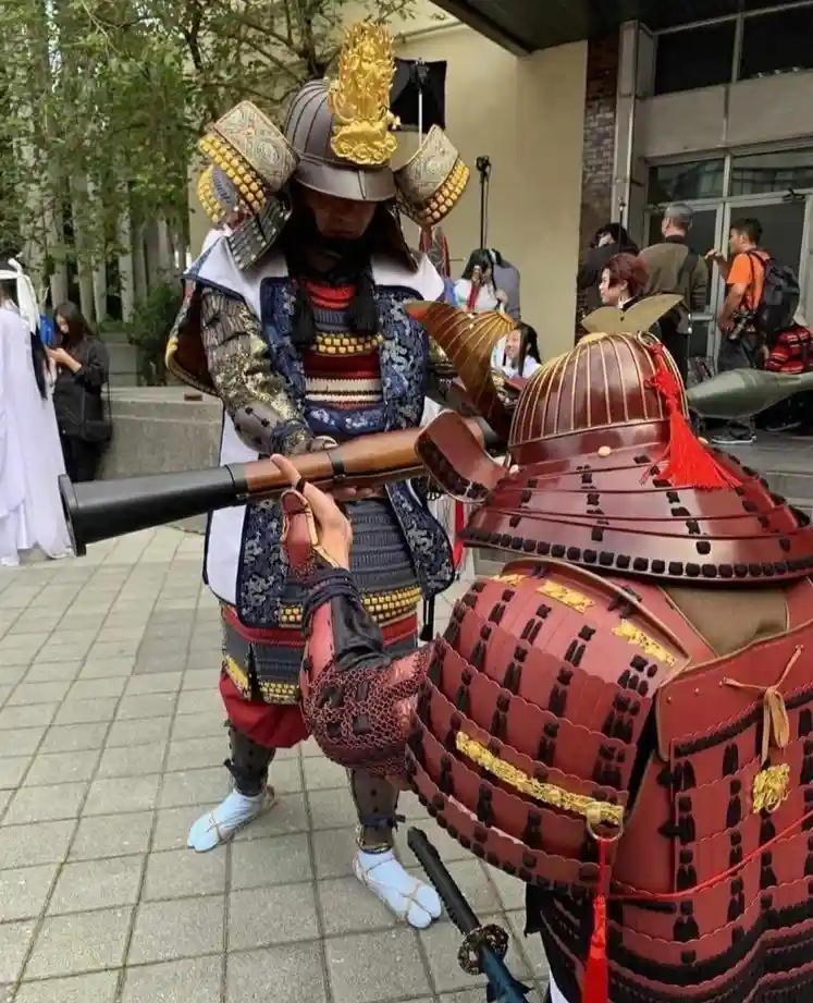 Two people dressed in elaborate, traditional Japanese samurai armor stand outdoors on a paved area. The samurai in the…