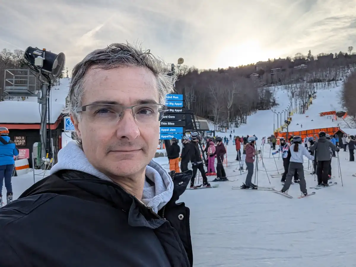 A man with glasses and a black jacket takes a selfie at a ski resort, looking slightly unenthusiastic. Behind him, skiers…