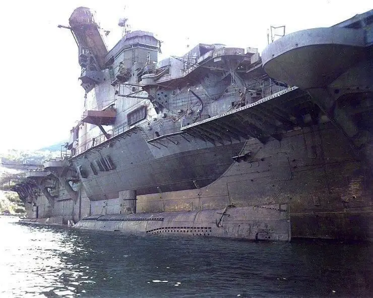 A large, weathered aircraft carrier is shown docked, with a submarine visibly wedged underneath its hull, creating a comical…