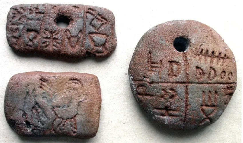 Three ancient, weathered clay tokens with carved symbols and holes, one of which clearly shows the text 'DIDOO' carved into…