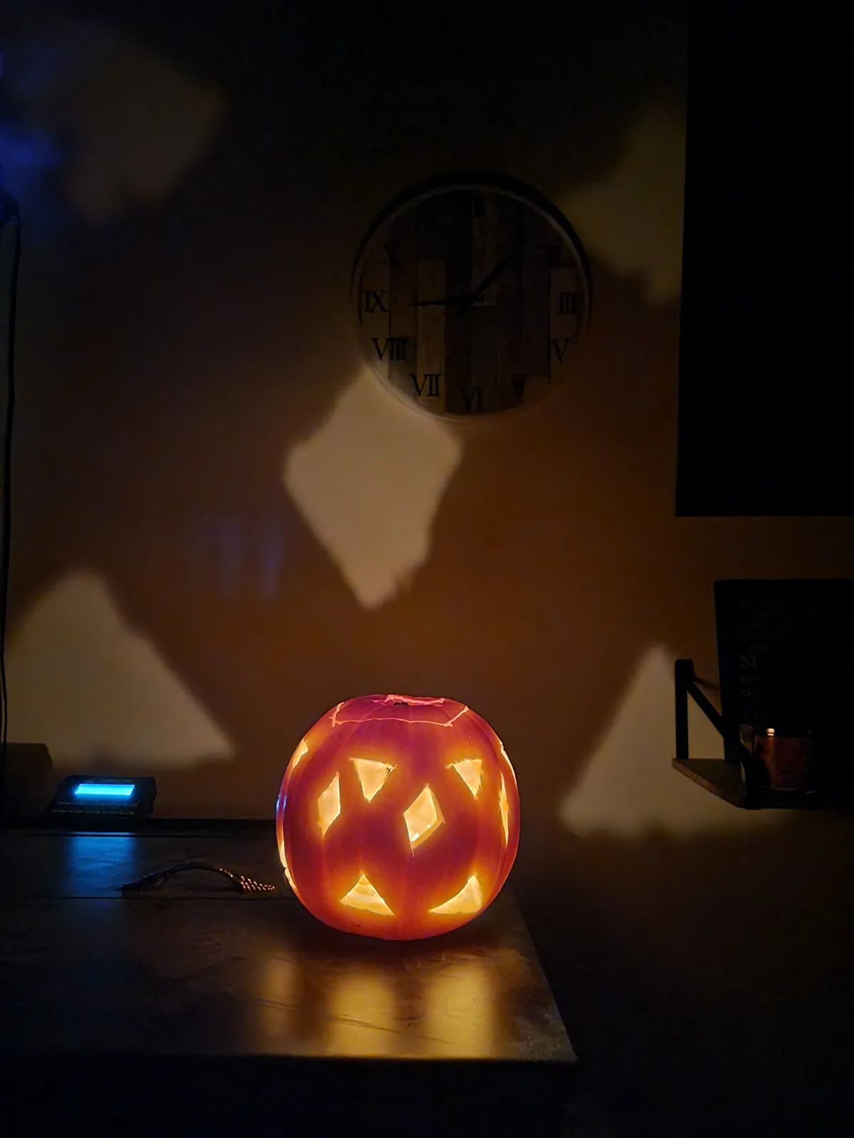 A carved jack-o'-lantern with a glowing orange light inside sits on a dark wooden surface, casting a warm glow. Behind it, a…