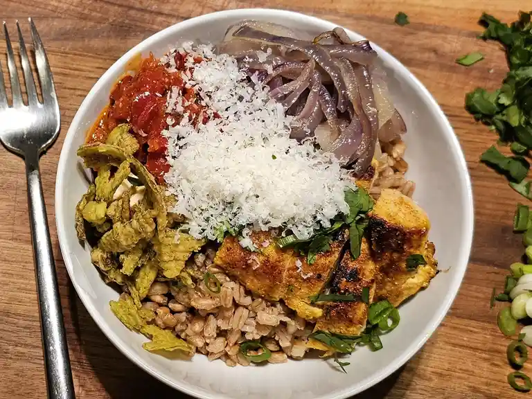A white bowl filled with brown rice, grilled tofu, roasted red peppers, shredded coconut, caramelized onions, chopped…