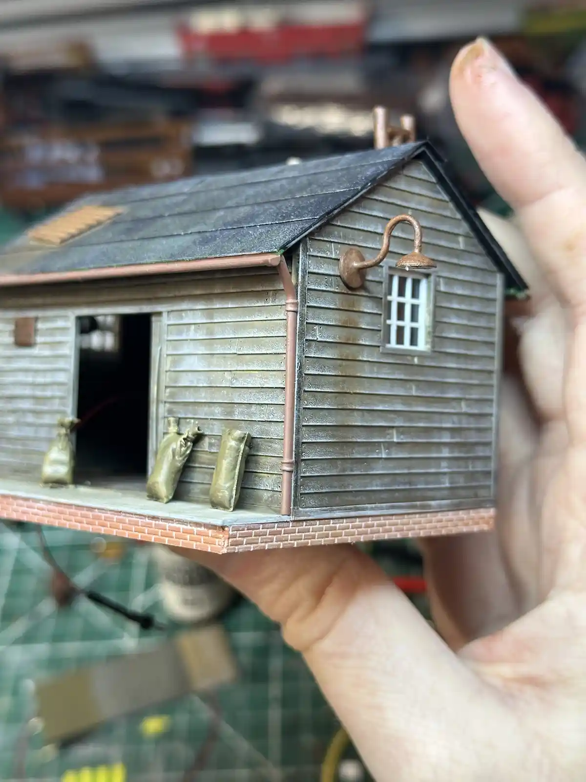 A hand holds a detailed miniature model of a weathered wooden house with a brick foundation, a window, and a shower head…