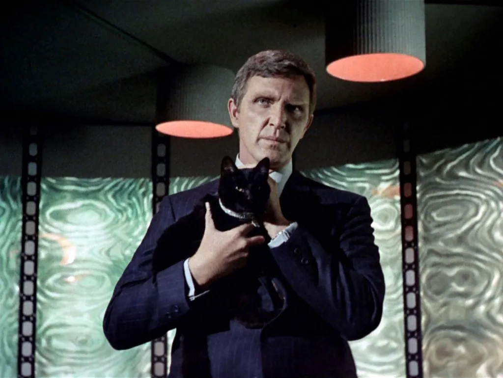 A man in a dark suit holds a black cat with a collar, looking sternly at the camera. The background features abstract green…
