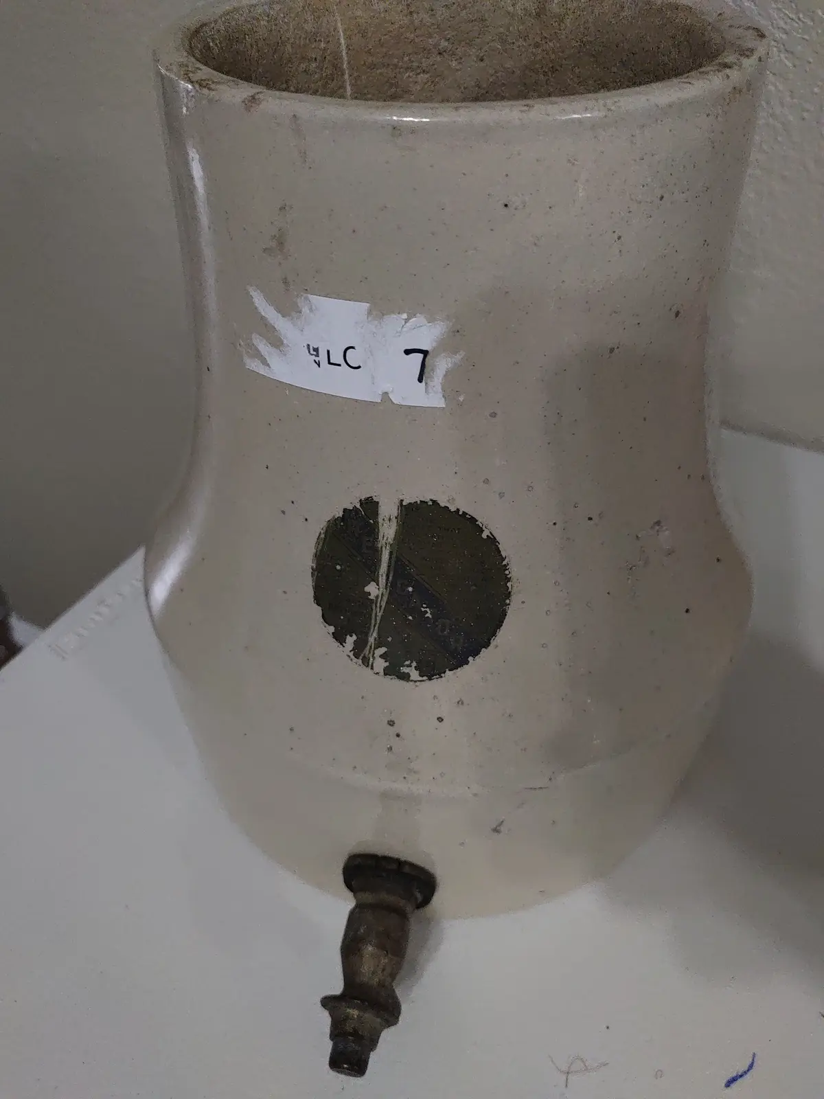 A close-up of a beige ceramic water cooler with a metal spigot at the bottom, a torn white label reading 'LC 7' on its side…