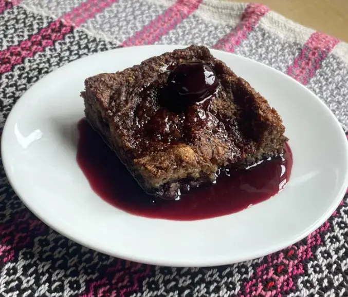 A square piece of dense, dark brown cake sits on a white plate, generously topped with a glossy, dark red sauce that pools…