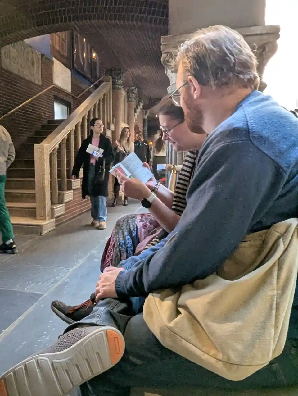 A man in a blue sweater and glasses sits on a stone bench outdoors, leaning over a woman who is holding a booklet or…