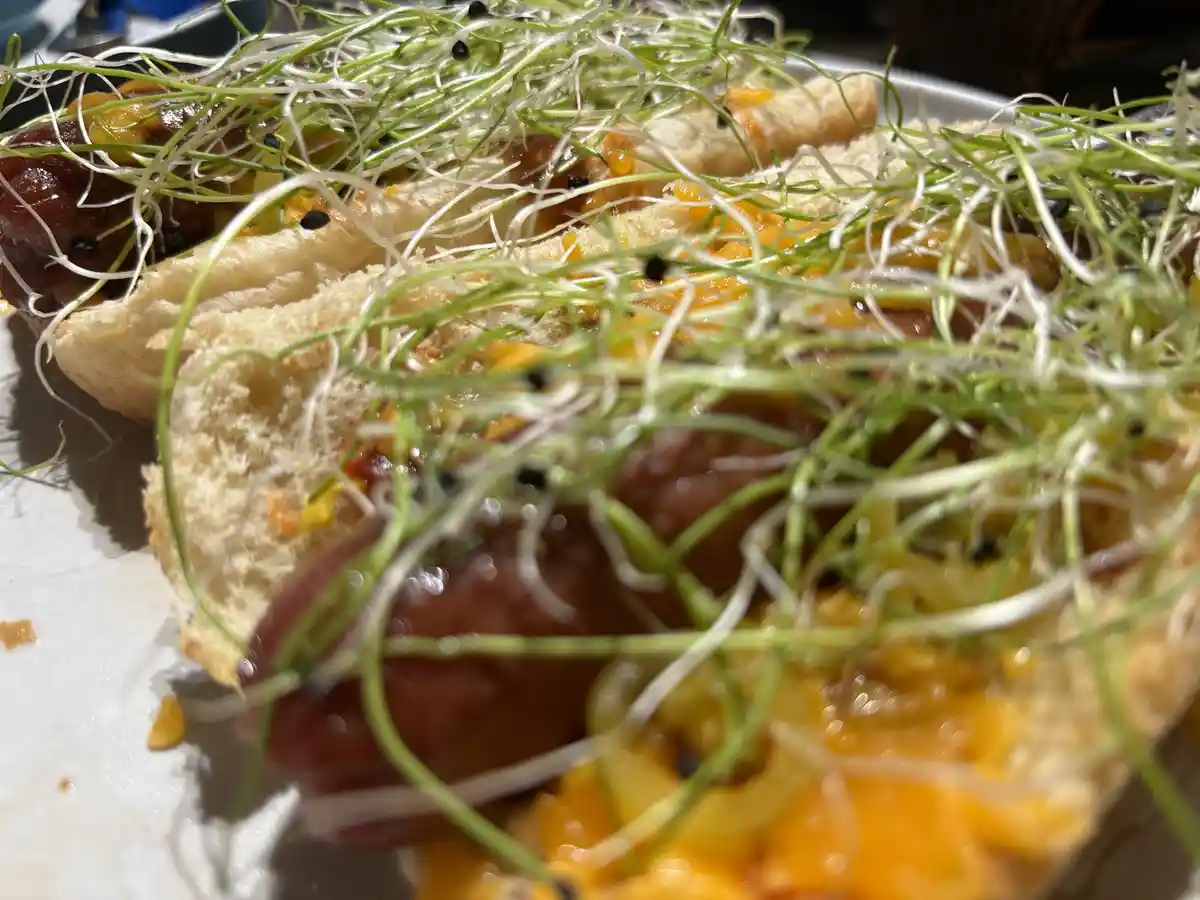 A close-up photo shows two hot dogs in buns, generously topped with shredded orange cheese, a red sauce, and a large amount…