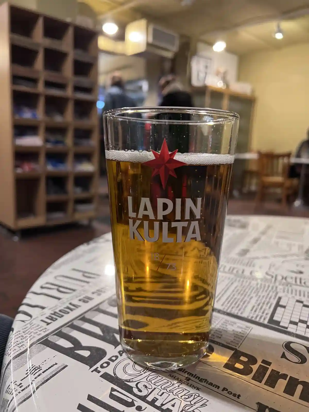 A pint glass filled with golden beer, branded with 'LAPIN KULTA' and a red star logo, sits on a table covered with newspaper…