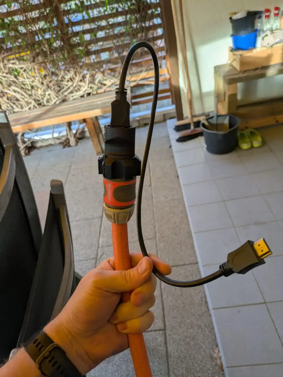 A person holds an orange garden hose with a black garden hose nozzle attached to its end. A black HDMI cable is plugged into…