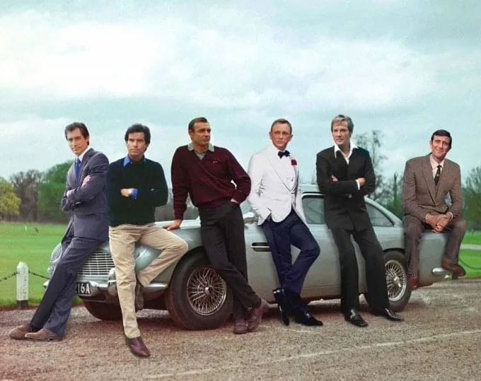 six men in various suits and casual attire are posing confidently around a silver Aston Martin DB5 on a gravel road, with a…