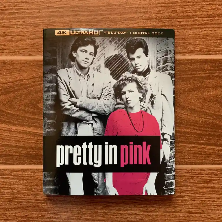 A 4K Ultra HD Blu-ray case for the movie 'Pretty in Pink' rests on a wooden surface. The cover art features a…