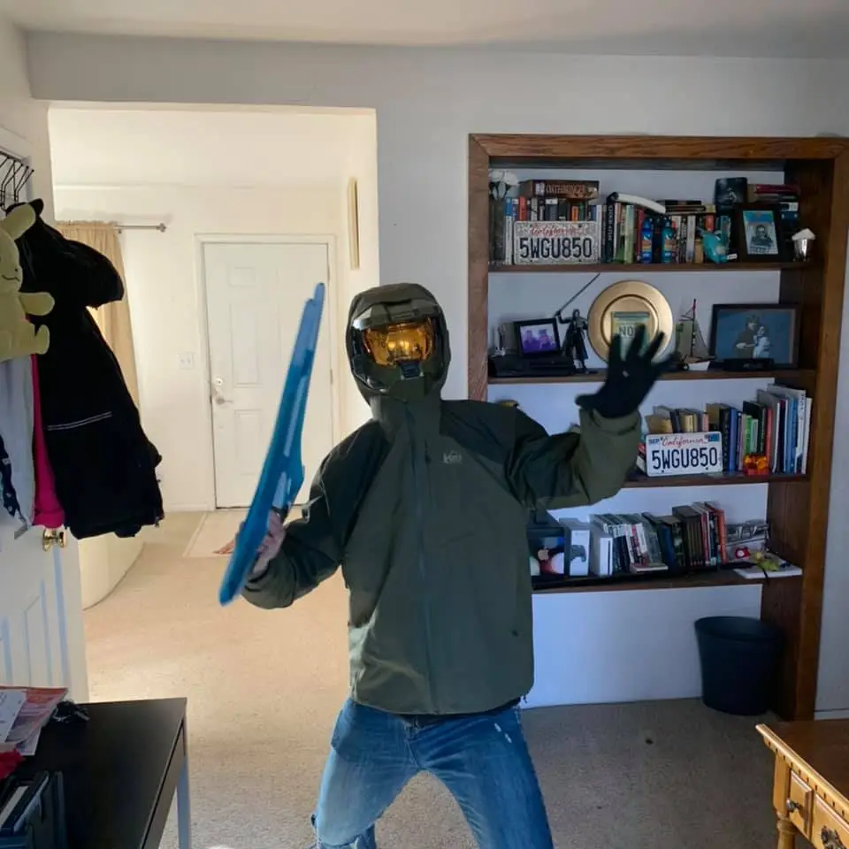 A person wearing a green Halo Master Chief helmet and jacket stands in a living room, holding a blue plastic sword in one…