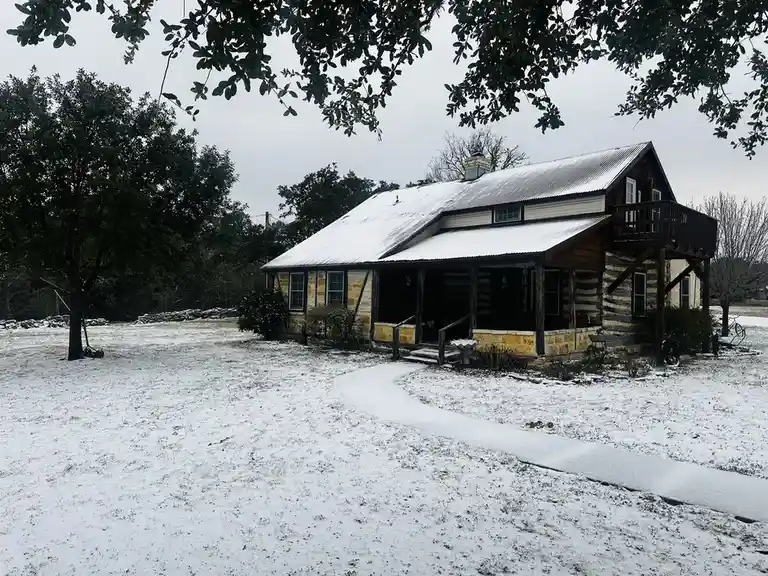 A rustic log cabin with stone accents and a metal roof is covered in snow, with snow also blanketing the yard and a winding…