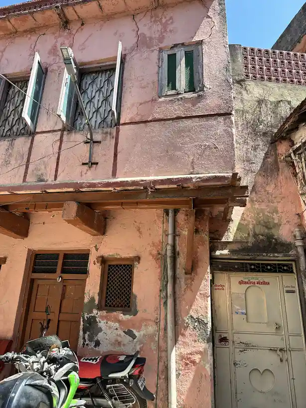 A photo of a weathered, pinkish-orange building with cracked walls, wooden beams, and windows with metal grilles. A…