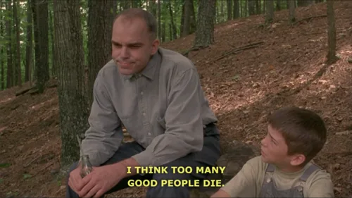 A still from a movie shows a man sitting in a forest with a young boy beside him. The boy looks up at the man, and yellow…