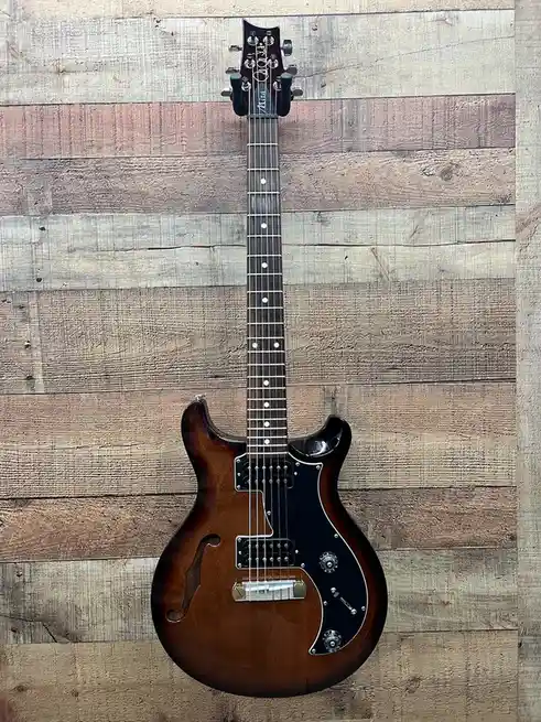 A sunburst-finished electric guitar with a black pickguard and white dot inlays is photographed vertically against a rustic…
