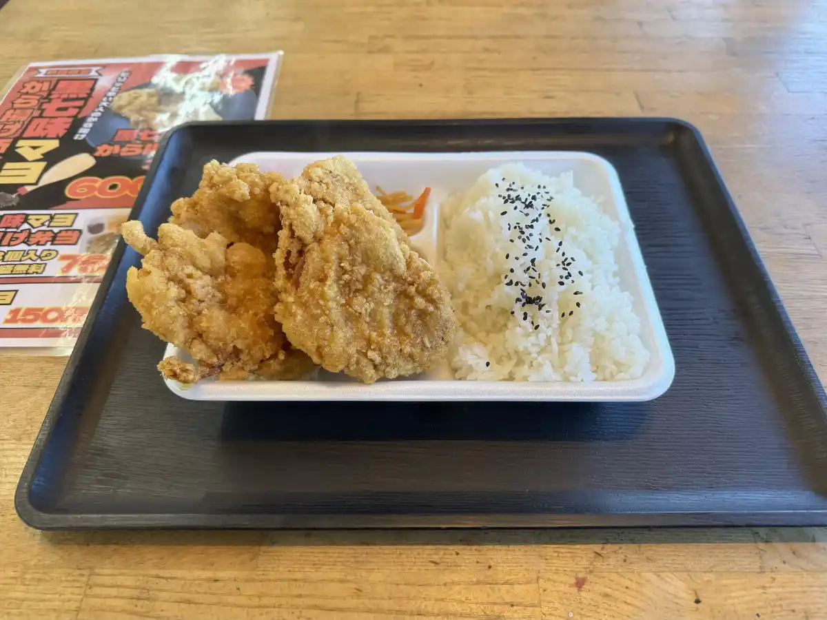 A white styrofoam bento box on a black tray contains two large pieces of golden-brown fried chicken, a small portion of…