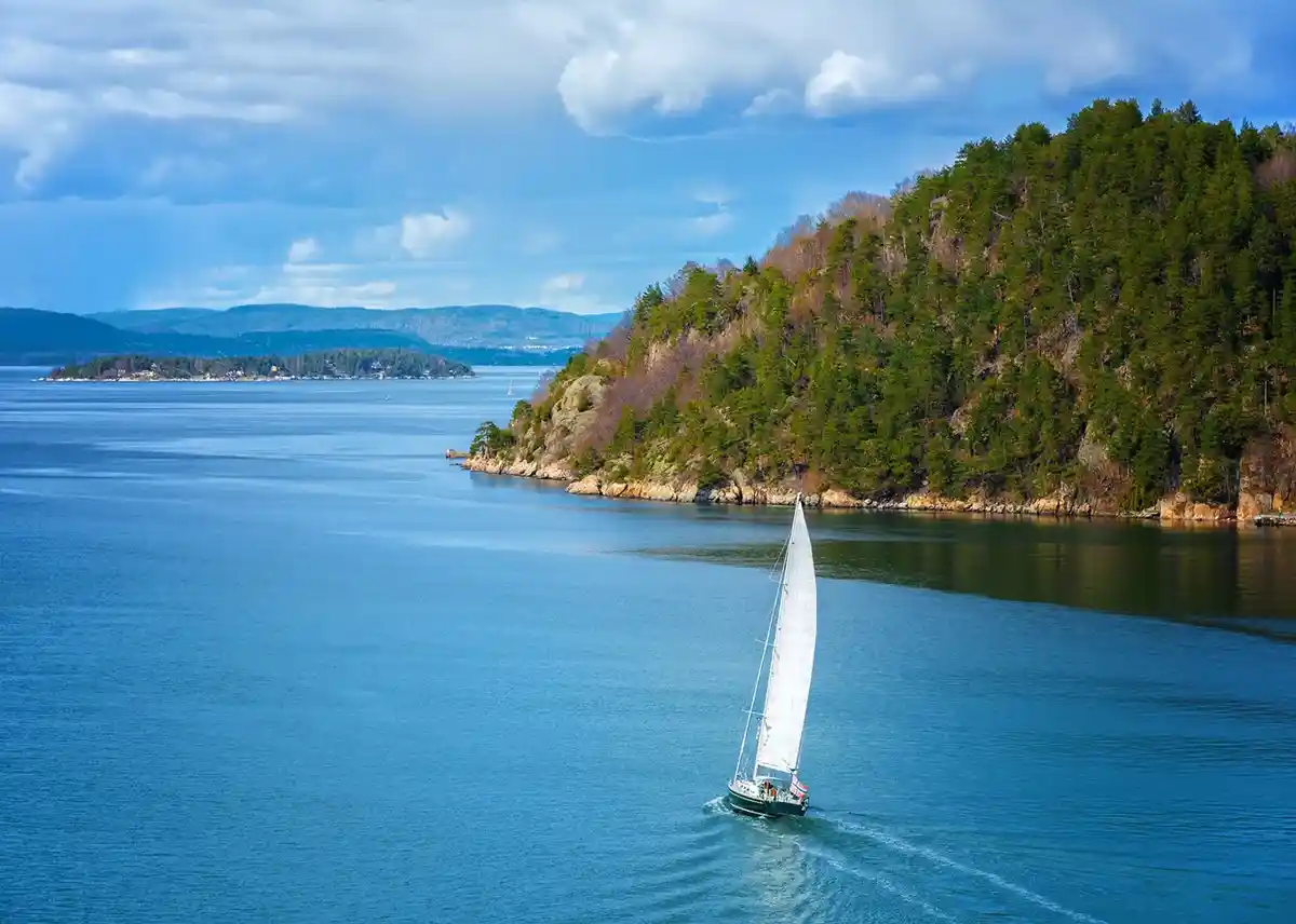 A serene landscape photo shows a white sailboat gliding through calm blue water beside a forested, rocky shoreline under a…