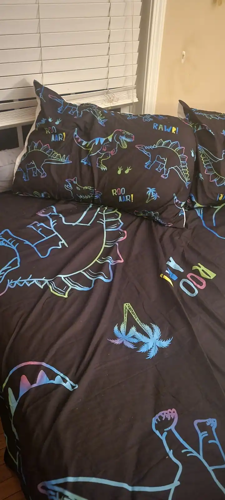 A close-up photo of a dark brown bed with a dinosaur-themed comforter and pillows, featuring colorful outlines of various…