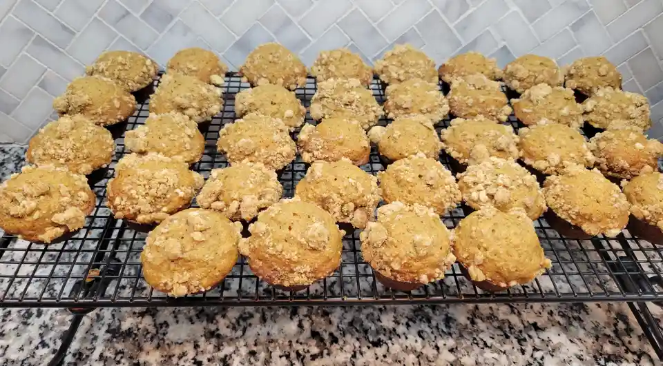 A large batch of golden-brown muffins with crumbly streusel toppings sits neatly arranged on a black wire cooling rack. The…