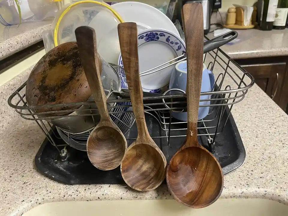 A close-up photo of three wooden spoons hanging from a metal dish rack filled with dirty dishes, including a heavily stained…