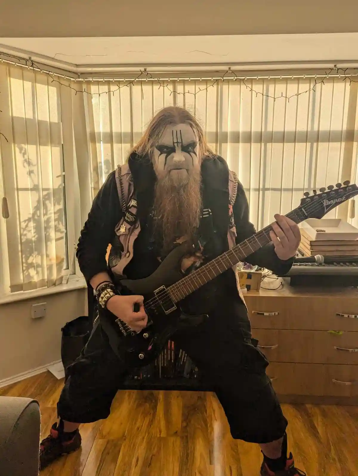A person with long hair, a long beard, and heavy black-and-white face paint resembling a metal musician is posing with an…