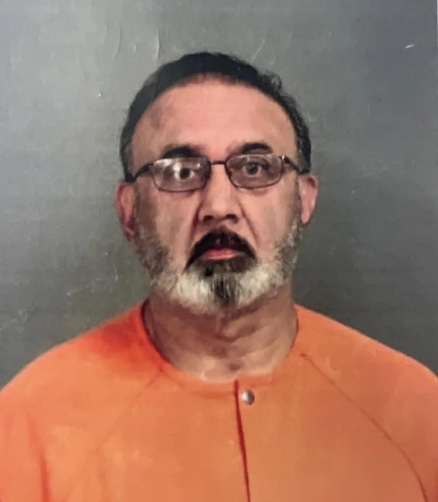A mugshot-style photo of a middle-aged man with glasses and a graying beard, wearing an orange jumpsuit against a plain gray…