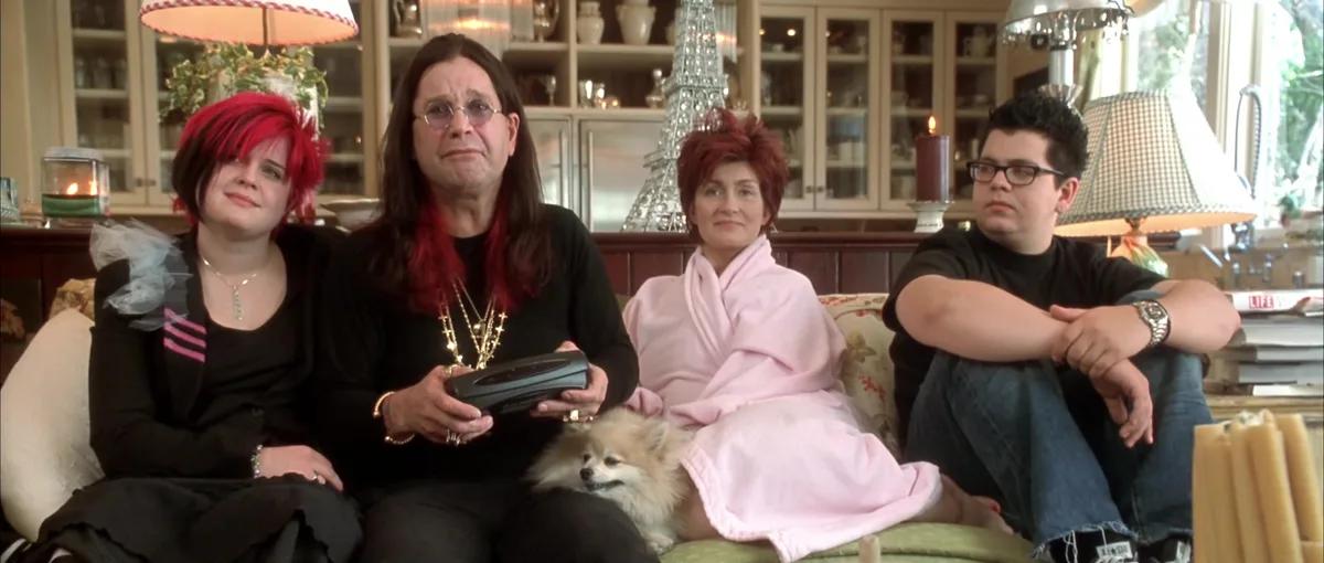 Four people sit on a couch in a living room, looking bored or unimpressed. The person in the center holds a remote, while a…