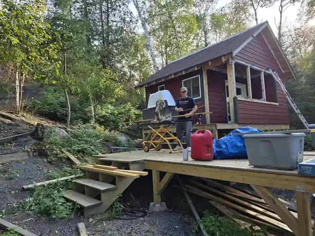 A man in a cap and t-shirt stands on a wooden deck in front of a partially built red cabin, holding a tape measure and a…