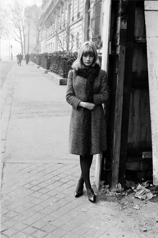A black-and-white photograph of a woman with blonde hair standing on a sidewalk, leaning against a wooden structure. She…