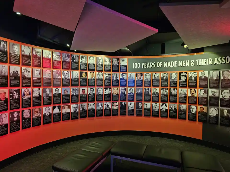 A curved museum wall displays dozens of black-and-white portraits of men, each with a biographical text panel, under a large…