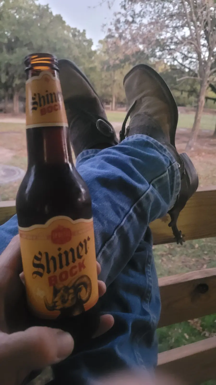 A person wearing blue jeans and dark cowboy boots has their feet propped up on a wooden railing outdoors, holding a bottle…