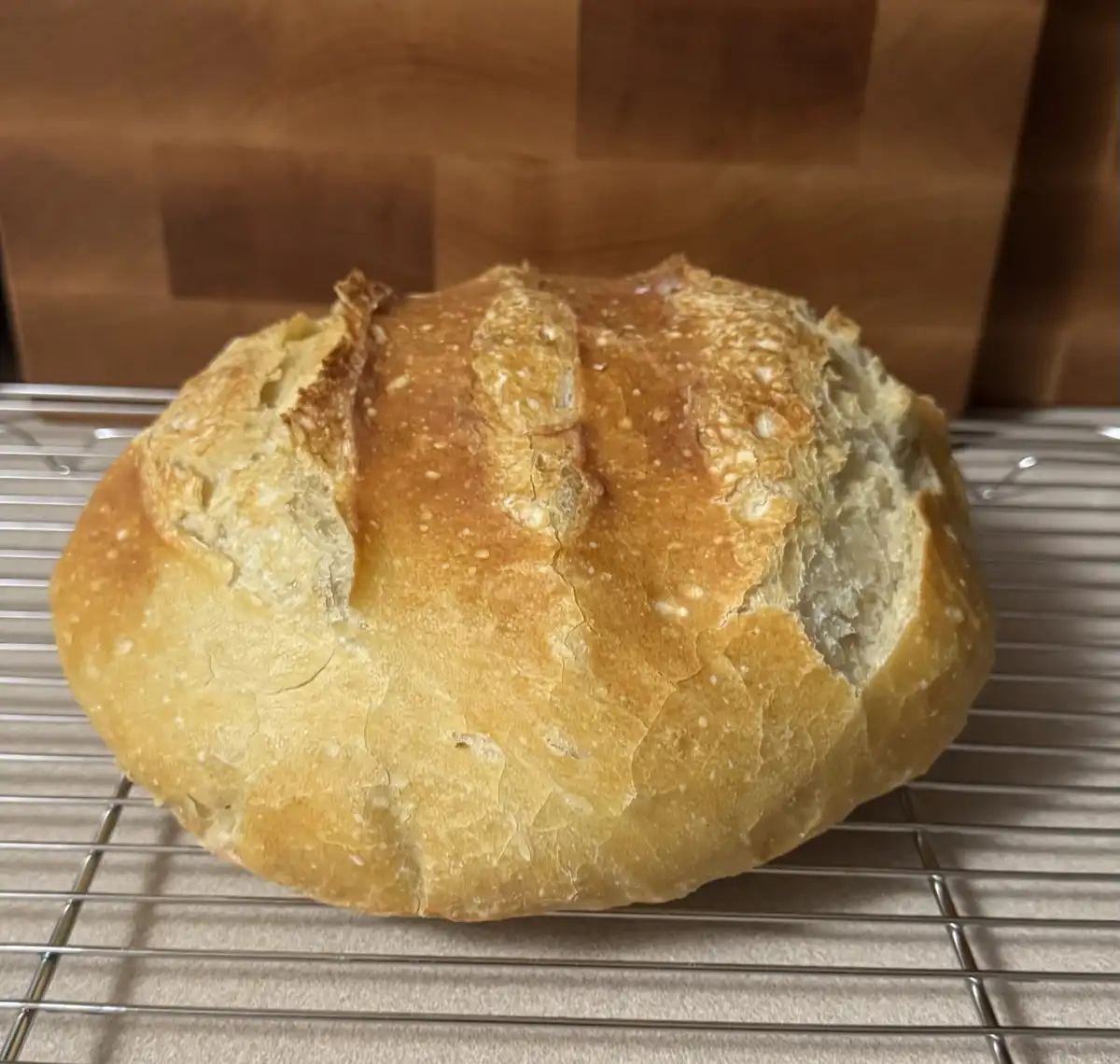 A round, golden-brown loaf of artisan bread with a cracked crust sits on a metal cooling rack, with a wooden surface in the…