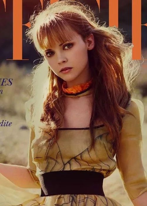 A woman with long brown hair and bangs stares directly at the camera with a serious expression, wearing a yellow dress with…