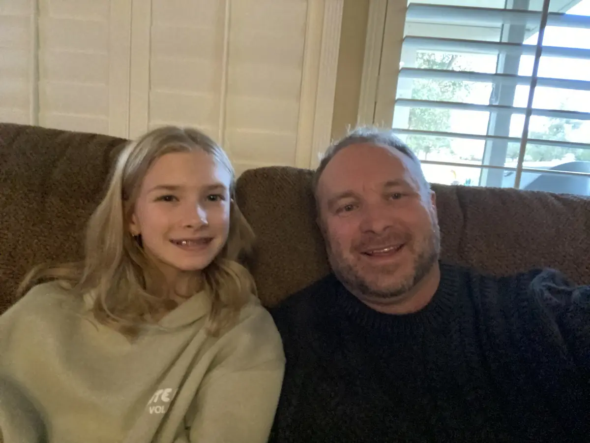 A man and a young girl are sitting side-by-side on a brown couch, smiling at the camera. The man, on the right, wears a dark…