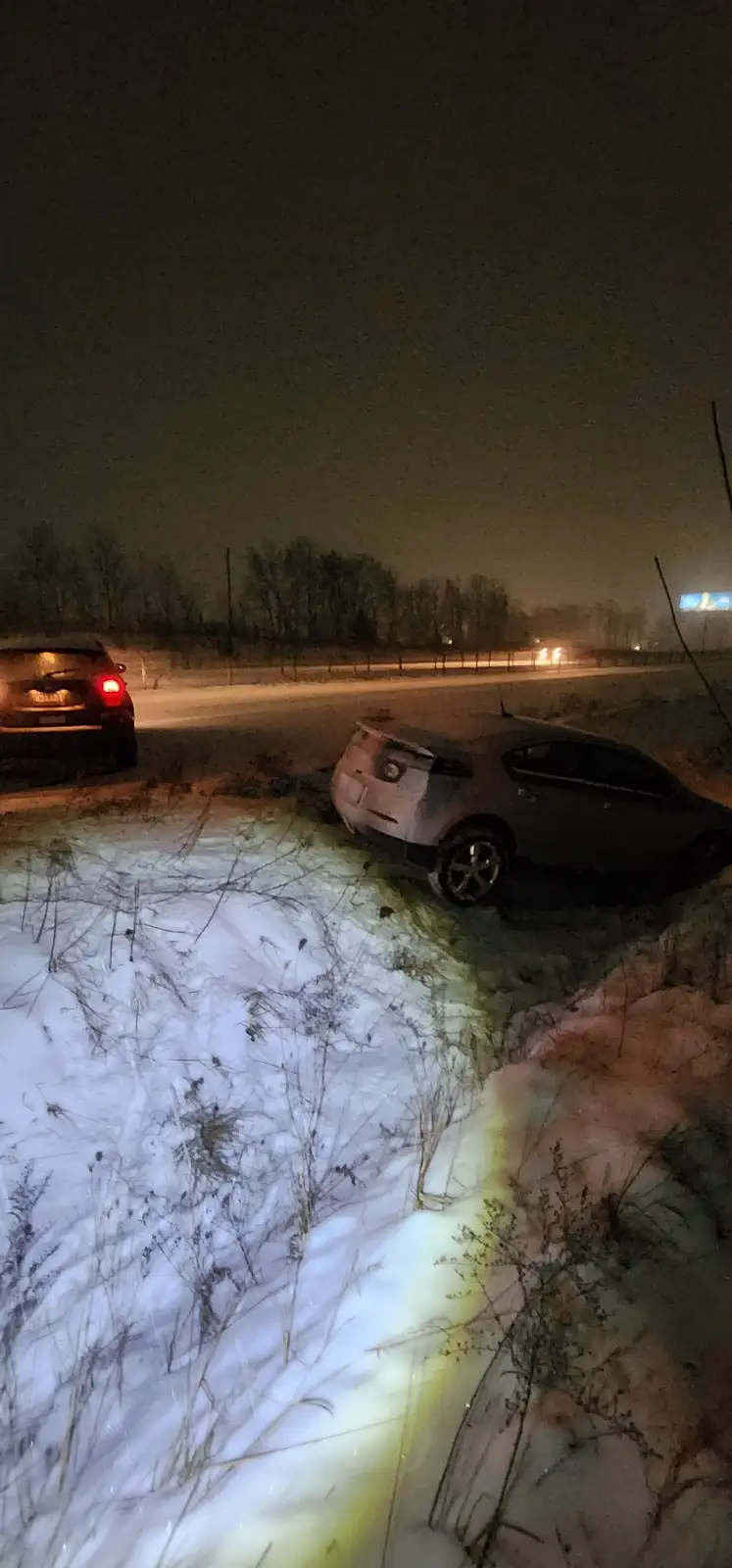 A dark-colored car is stuck sideways in a snowy ditch at night, illuminated by a bright yellow flashlight beam on the snow…