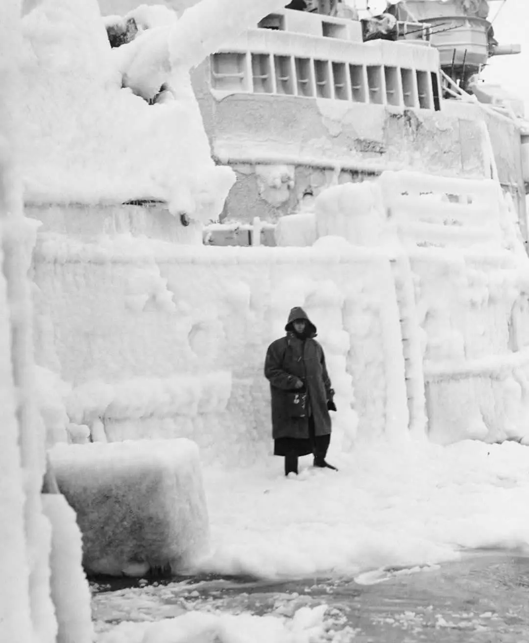 A black-and-white historical photo shows a person in a heavy winter coat and hood standing in front of a massive ship…