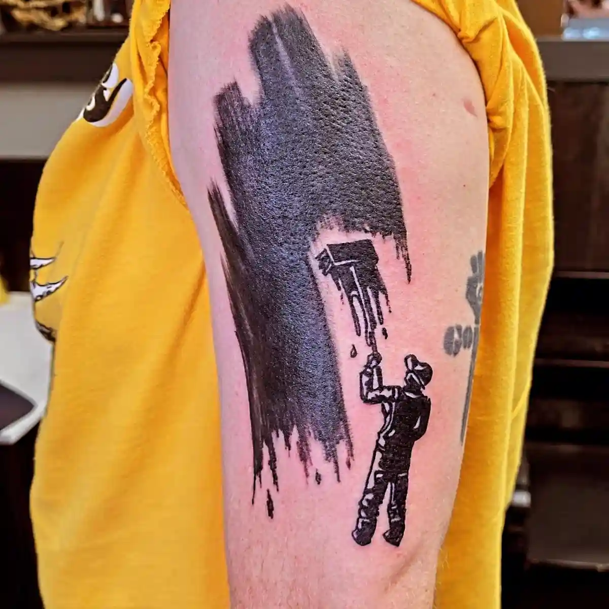 A person's upper arm displays a black ink tattoo of a small figure holding a paint roller, painting a large, dripping black…