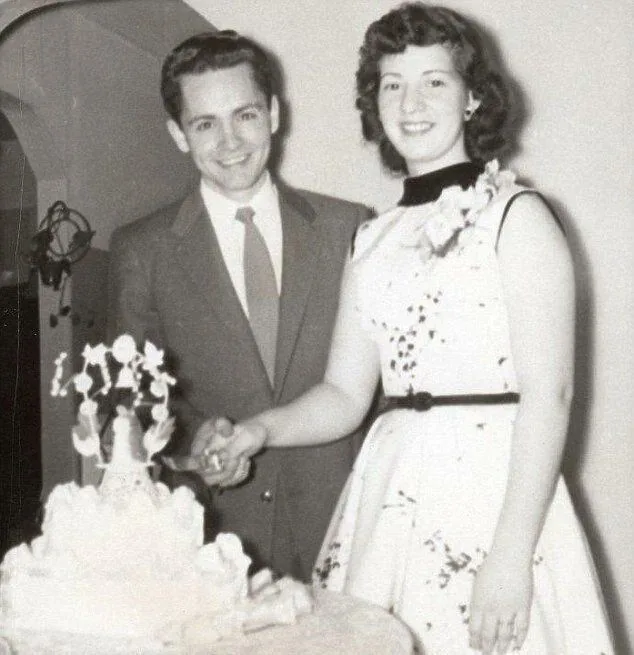 A black-and-white photo of a smiling couple in vintage wedding attire standing beside a large, ornate wedding cake. The…