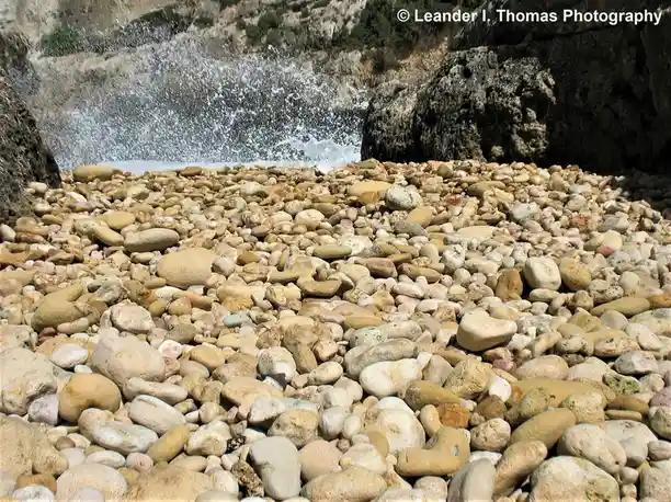 A photo of a rocky beach with water crashing against rocks in the background. The foreground is filled with smooth, rounded…