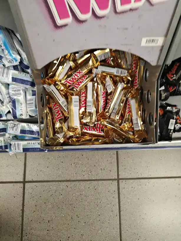 A close-up shot of a supermarket shelf displaying a bin overflowing with individually wrapped Twix chocolate bars in their…